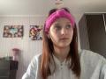 AdrianneEllinger with pink knit headband, gray hoodie, and white earbuds, has brown hair, light skin, and is looking to the side with a neutral expression.