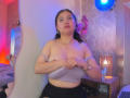 CristineBernard with medium tits, pulling down a gray bra, sitting in a room with blue and red lights, black skirt, and a small lamp on a wooden table.