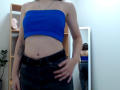 Steisii with blue tube top, showing her tits and flat stomach, wearing black pants with a belt, her hand on her hip, reflected in a mirror.