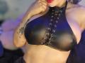 Kkole's big tits in a black leather halter top with lace-up detail, showing a tattoo of a leaf on her right arm, and red lipstick.