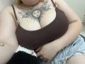 DiLaria has large tits with a sun and face tattoo above her cleavage, wearing a brown tank top and ripped jeans, sitting with a black and white fuzzy blanket.