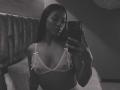 Amberv with dark skin and long hair takes a selfie in a mirror, wearing a sheer white bra with small bows, showing her small tits and nipples.