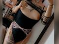 DenyChantal with exposed tits, wearing a black fishnet top, pink lace garter, and black bodysuit, showing tattoos, long nails, and a pearl necklace in a mirror selfie.