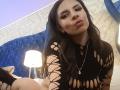 VioletaStone with long black hair, pouty lips, wearing a black, cut-out, mesh dress, and a silver necklace with a star, sitting on a bed with a lamp.