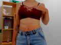 Steisii in red lace bralette, showing tits with cross-strap, wearing blue jeans, hand on chest, tattoo on left arm, standing in room with wooden shelf and plants.