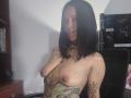 AmelieBlake with black hair, nose ring, and tattoos, topless, showing her small tits and tattoos, including a large green and black tattoo on her chest.