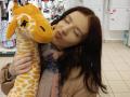 EviaMarcot kissing a yellow and white giraffe stuffed animal, eyes closed, pink and blue scarf, brown coat, brown hair, white tiled floor, white shelves in background.