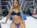 CloeRouss with blonde hair, blue lace bra, and matching panties, sitting on a bed, showing her tits and pussy, with a slight tan and red lipstick.