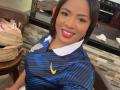 AliceBooty with medium brown skin, black hair, pink lipstick, blue striped sports jersey, white collar, yellow Nike logo, smiling, sitting at a bar, bag on wooden chair.