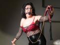 KarlaHartley with black fishnet sleeves, red bra with exposed tits, black leather pants, ball gag in mouth, holding red and black leather flog, dark hair, glasses, serious expression.