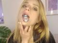 LyannaDifran with blonde hair, brown eyes, black shirt, and pink gem butt plug in her mouth, hand near chin, looking up. Pupils visible through gem.