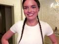 DrusillaMaiava, with braids, white shirt, and smiling, has small tits, a cute face, and a pierced chin, standing in a room with a chandelier.
