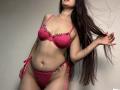 Tisia with long brown hair, wearing pink lace bra and matching panties, holding hair to the side, showing her tits and pussy, standing against a plain wall.