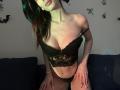 Valnes, con lencería negra de encaje, mostrando sus pechos y vulva, inclinada hacia adelante sobre un sofá oscuro, con su cabello teñido de verde cayendo hacia un lado, y las manos sobre sus muslos..