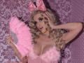 MademoiselleBella with blonde curls, pink bow, and fluffy pink fan, holding it near her tits, wearing a pink feathered top, and a diamond necklace, looking sexy.