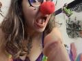 ArianaBlonde with a red nose, black tattoos, and glasses, sticking out her tongue, holding a flaccid, uncircumcised cock, wearing a purple lace bra.