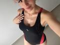 EliseMonet in a black and red one-piece swimsuit, shiny with sweat, showing her boobs, pink hair, and a cute smile, hand on chest, white nails, in a selfie.