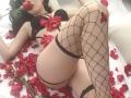 IvannaSinn lies on a white surface, wearing black lingerie and fishnet stockings, with red rose petals covering her body. Her legs are crossed, cock exposed, black hair, pale skin.