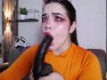 AryButty with dark hair, orange shirt, and pink eye makeup, fellating a large, black, dildo. Her mouth is wide open, lips wrapped around the dildo's thick, veiny head.