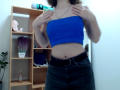 Steisii with blue tube top, hands on chest, black high-waisted skirt, showing a bit of her belly, standing in a room with a wooden shelf and green plant.