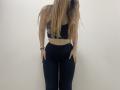Palerianna stands against a plain white wall, facing forward. She has long, straight brown hair covering her face. She wears a black sports bra with a subtle pattern and black high-waist leggings. Her right hand rests on her hip while her left hand is relaxed by her side. She has a black bracelet on her right wrist. The image focuses on her midsection and lower body, with a minimalist composition. Her skin is light. The overall style is simple and modern.