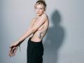 Topless EolantaMarina with blue eye makeup, black pants, holding chain, tattoo on right side, pale skin, blonde hair in messy bun, shadow on gray wall, arms crossed.
