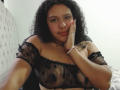 AriaVega with curly black hair, wearing a sheer black off-shoulder top, resting her hand on her cheek, showing her tits, smiling slightly, in a bedroom.
