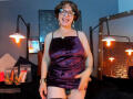 SamantaMature69 with short brown hair, glasses, purple velvet dress, exposing a black butt plug, smiling, standing in a room with hanging lights and a brick wall.