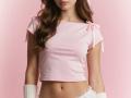 Diantrisy with long brown wavy hair, pink ribbed crop top tied on shoulders, white ribbed wrist warmers, pink background, showing her flat stomach and small tits.
