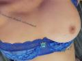 De borsten van FrenchShanna, in een blauwe kanten beha, met een kleine turquoise hanger, waarbij een tatoeage en een zichtbare tepel te zien zijn. Haar huid is licht..