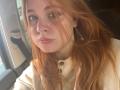 ClemenciaGerhart with red hair, freckles, and blue eyes, wearing a beige button-up shirt, has a small bruise near her right eye, and a heart-shaped earring.