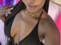VenusWilly with dark skin, glossy lips, and braided hair wearing a black halter top showing her large, shiny tits. She's also got a delicate necklace with small pearls.