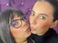 Close-up photo of two women, ValkiriaAndDione on the left with black hair, glasses, and a choker, kissing ValkiriaAndDione on the right with dark hair and pink lips. Purple background. Sexual tension.