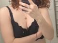 ChayaParrett's tits in a black lace bra, holding a phone with a ring on her finger, curly red hair, wearing a necklace and bracelet, in a bathroom mirror selfie.
