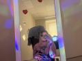DiLaria with blonde hair, holding phone in front of mirror, wearing shiny black dress, showing tits with large black tattoo on chest, standing in room with colorful lights.
