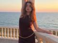 LeScharwath with wavy brown hair, wearing a black dress and gold chain, arms raised, leaning on a white railing at sunset, ocean in background, standing on a stone balcony.