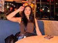 JennTylor with long black hair, brown tank top, blue jeans, black belt, black jacket, and black bag, sitting at a wooden table, hand in hair, phone on table.