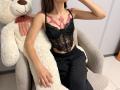 AmeliaMaiv with pink tattoo on chest, wearing black lace corset and black pants, sitting on large white stuffed teddy, hand near face. Boobs visible through lace.