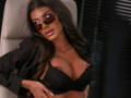 DariaAmaris with dark skin, long brown hair, and brown sunglasses, wearing a black lace bra that shows off her large, shiny, bare tits, leaning back in a chair.