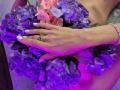 TenerliRize's hand with white nails and silver ring rests on purple and pink hydrangea flowers, pink bracelet visible, under purple lighting.