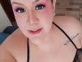 GiiannaMorrys with pink eyeshadow, black eyeliner, nose piercing, septum ring, black mesh top, tattoo on right shoulder, looking up, light skin, pink blush, small earrings, wood floor background.