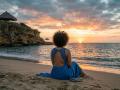 RoldanStefy with curly hair, blue backless dress, sitting on sandy beach, facing sunset, book on sand, rocky cliff with hut on left, orange and pink sky, ocean.