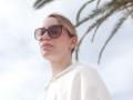 MissFrenchy with sunglasses, blonde hair, white shirt, and earrings, looking up with a slight smile, palm leaves in background, soft light, relaxed vibe.