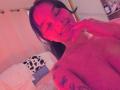 Yurimy, topless, smiling with eyes closed, pink-red lighting, dark hair, butterfly tattoos on right shoulder, hand near mouth, visible breasts, light skin, bedroom setting, relaxed, playful.