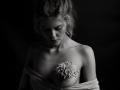 Black-and-white photo of EolantaMarina with wavy blonde hair, eyes down, holding a flower over her bare tits, wrapped in a towel, dark background.