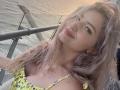 EliseMonet has wavy, pinkish-blonde hair, a cute smile, and is wearing a yellow, floral-patterned dress with a low neckline, showing her small tits. She's on a boat.