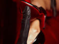 AfroditaDoir's shiny red high heel is on her black leather boot, with her bare foot showing. The heel is pointed upwards, showing the sole's texture.