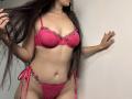 Tisia with long dark hair, wearing pink lace bra and panties with floral accents, holding her hair, showing her tits and pussy, standing against a white wall.