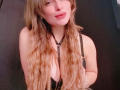 GynaHykarii with long wavy blonde hair, black leather harness, and cleavage showing, smiling with pink lips, arms behind her back, in a dimly lit setting.
