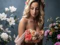 ViktoriiaLu with wavy blonde hair, wearing a strapless dress made of pink and peach roses, surrounded by white and pink flowers, looking downward with a serene expression.